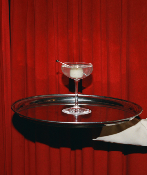 Cocktail transparent en coupe servi sur plateau argenté par un serveur en gants blancs, rideau rouge en fond – Cat Club