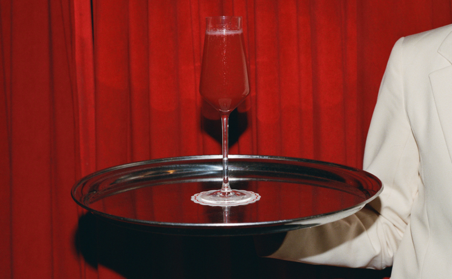 Cocktail rouge p&eacute;tillant en fl&ucirc;te pos&eacute; sur plateau argent&eacute; tenu par un serveur, rideau velours rouge en fond &ndash; Cat Club